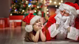 Image of a holiday photoshoot with small child and Santa. Christmas tree and presents in the background.